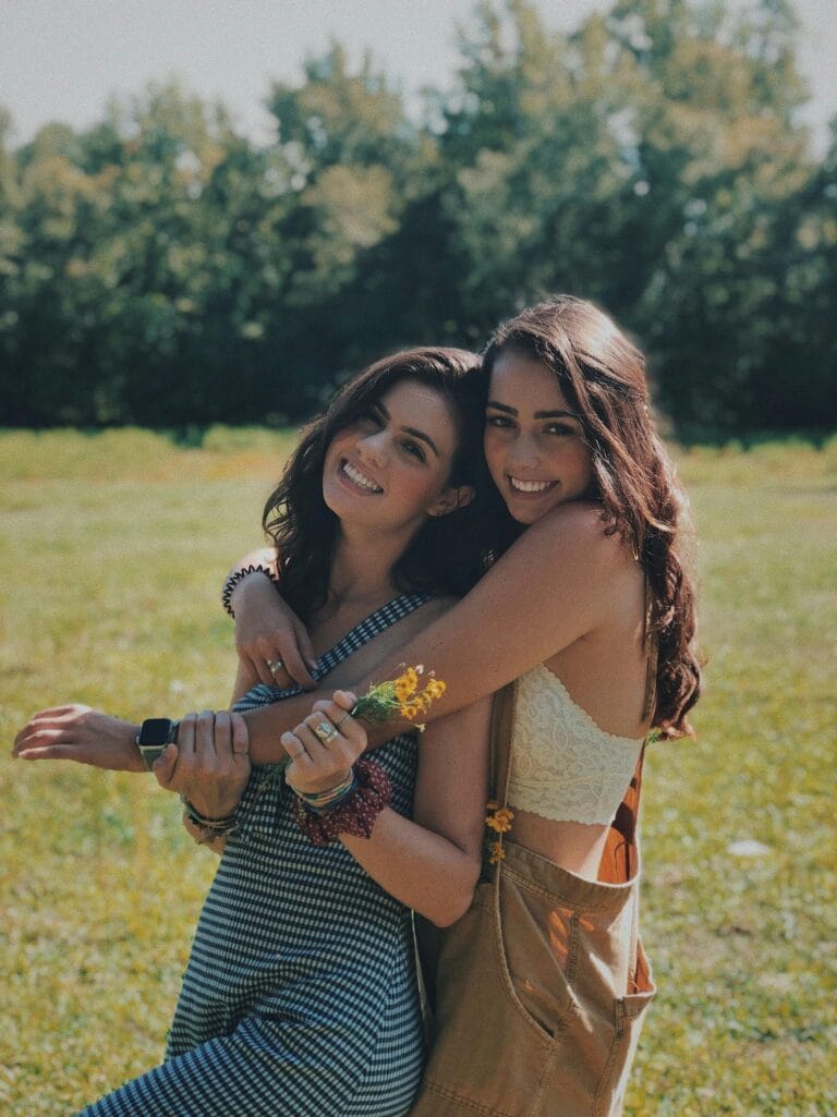 Two smiling sisters embracing in a sunlit park, enjoying a warm summer day together.