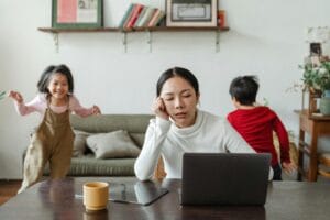 A tired mother working on a laptop while her children play around indoors